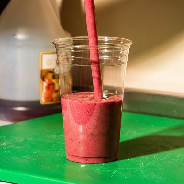 A vibrant pink smoothie is poured into a clear plastic cup on a green cutting board.